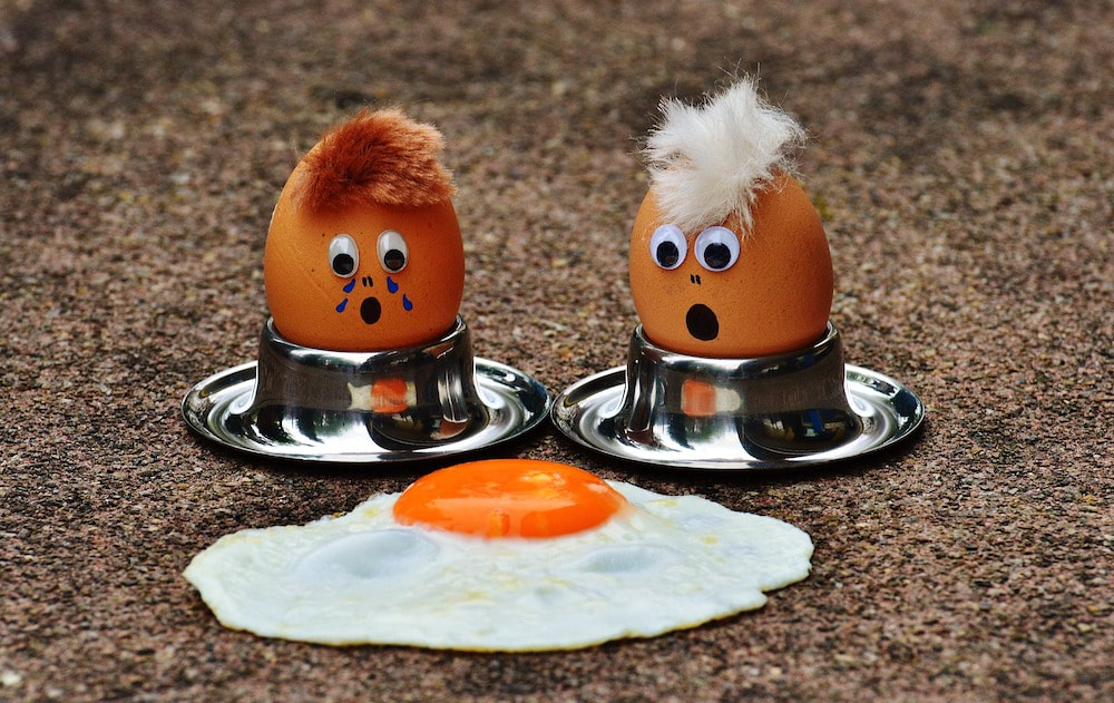 Two boiled eggs with faces staring in horror at a fried egg on the ground