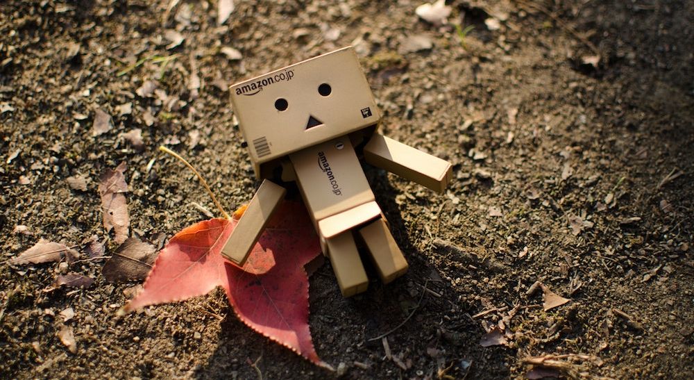 A robot made of tiny Amazon boxes lying alone on the bare ground.